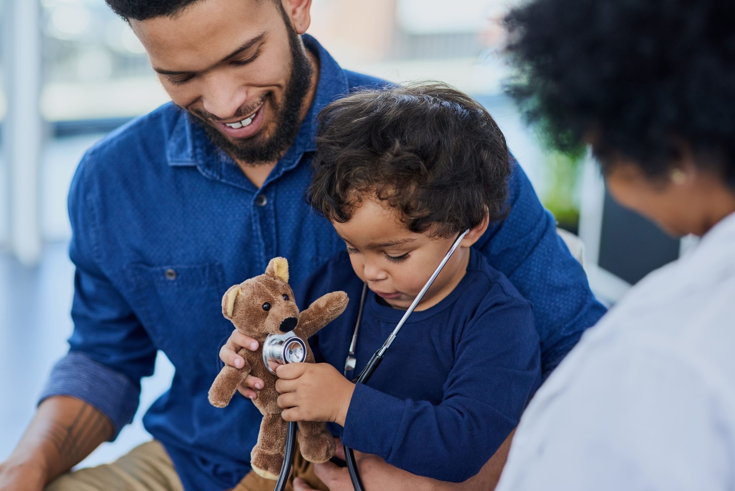A young child at the doctor's office sits on his smiling dad's lap and uses a stethoscope to listen to his small teddy bear. A doctor stands off to the side.
